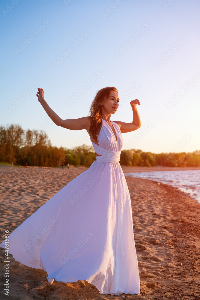 beautiful slender young woman of Caucasian ethnicity with long hair stands in a white dress in the wind near the sea at sunset