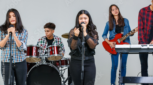 Group shot of teenage musicians playing the music and singing together on the weekends. Junior students play electric guitar, drum, and keyboard in the studio with the main vocalist singing in front