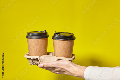 Hands holding two cups brown paper with black lid. Two coffee special offer or promo. Hands holding two cups on yellow screen background. Tea or Coffee to go. Brown paper cup with black lid.