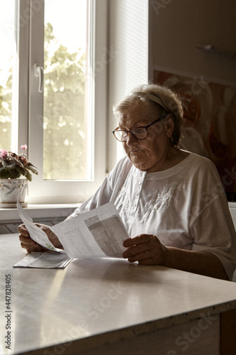 Elderly woman sitting at table in kitchen at home holds domestic bills, feels concerned forgot to pay or debt formed. Middle aged 80s retired woman
