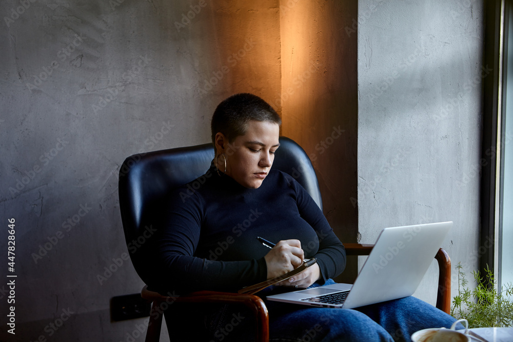 A young girl with glasses, with short hair is sitting in a cafe and working on a laptop. The girl looks at her laptop and makes notes in a notebook. The concept of distance learning A young girl with glasses, with short hair is sitting in a cafe and working on a laptop. The girl looks at her laptop and makes notes in a notebook. The concept of distance learning