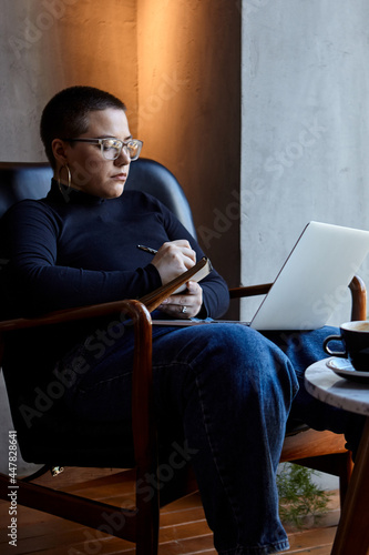 A young girl with glasses, with short hair is sitting in a cafe and working on a laptop. The girl looks at her laptop and makes notes in a notebook. The concept of distance learning