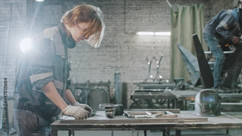 Young man with long hair working at the plant - brings metal long detail and instruction to the table and reading the instruction Young man with long hair working at the plant - brings metal long detail and instruction to the table and reading the instruction