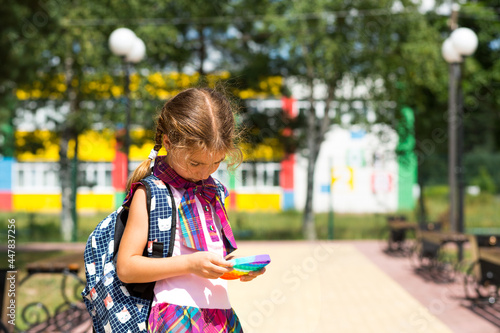 Little girl with a backpack and in a school uniform in the school yard plays pop it toy. Back to school, September 1. The pupil relaxes after lessons. Primary education, elementary class for student