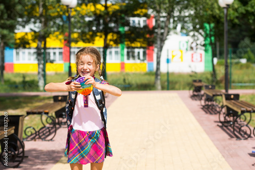 Little girl with a backpack and in a school uniform in the school yard plays pop it toy. Back to school, September 1. The pupil relaxes after lessons. Primary education, elementary class for student photo