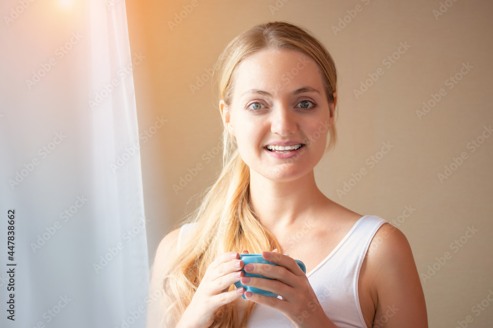 Beautiful young woman drinking coffee in the morning. Charming beautiful young woman hands holding hot cup of coffee or tea standing by the window in the bedroom. Beautiful young woman drinking coffee in the morning. Charming beautiful young woman hands holding hot cup of coffee or tea standing by the window in the bedroom.
