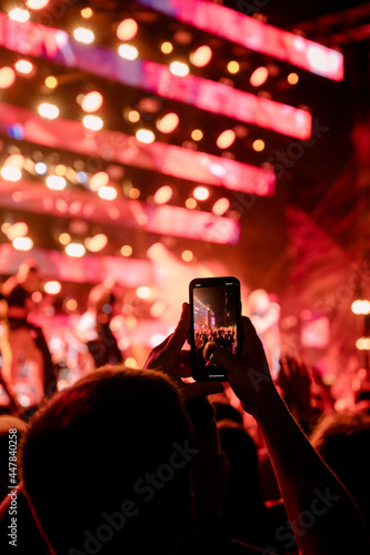 Music fan silhouette taking a photograph with a smartphone of a blurred musician at a concert with two hands. Red ambient light show. Video taking person.