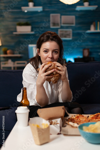 Portrait of caucasian woman looking into camera relaxing on sofa in living room enjoying takeaway food home delivery. Woman eating tasty delicious burger. Fastfood lunch meal order