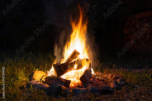 A colorful flaming bonfire with logs burning in the night. Fireplace with blazing firewood on the grass fenced with stones