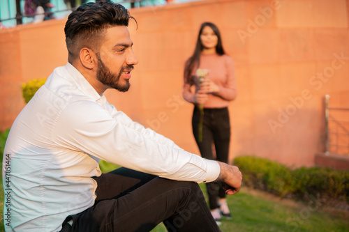 Happy young man sitting in the park and waiting for his girlfriend who comes with a red rose in hand.