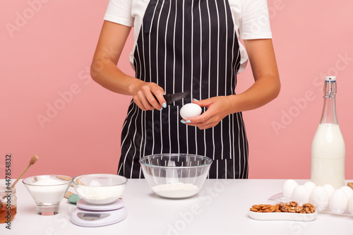 Cropped image of faceless woman baker doing pastry  driving egg to flour for making dough  wearing black striped apron  ingredients for bakery. Indoor studio shot isolated on pink background.