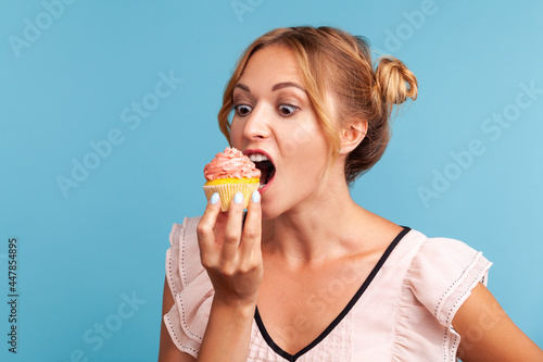 Sugar addiction. Portrait of hungry blonde young adult woman in dress biting delicious cake  looking with desire to eat sweet dessert. Indoor studio shot isolated on blue background.