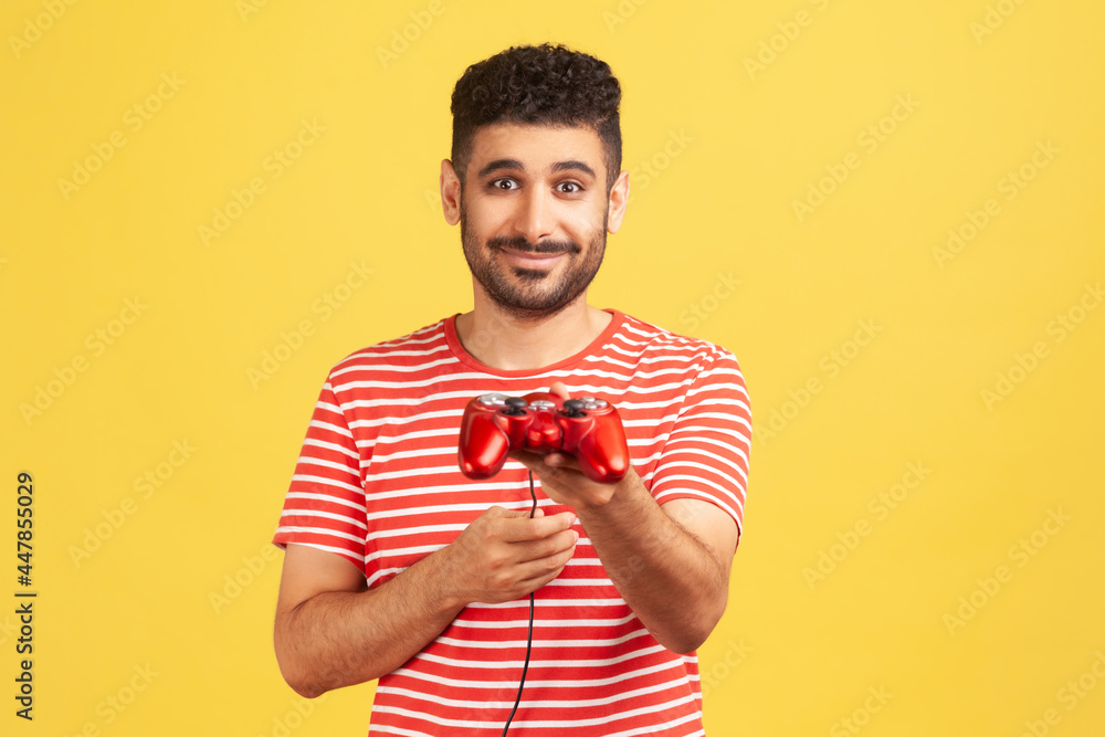 Generous bearded man in striped t-shirt holding in hand gaming joystick, suggesting you to play video game with him, virtual reality simulator. Indoor studio shot isolated on yellow background Generous bearded man in striped t-shirt holding in hand gaming joystick, suggesting you to play video game with him, virtual reality simulator. Indoor studio shot isolated on yellow background