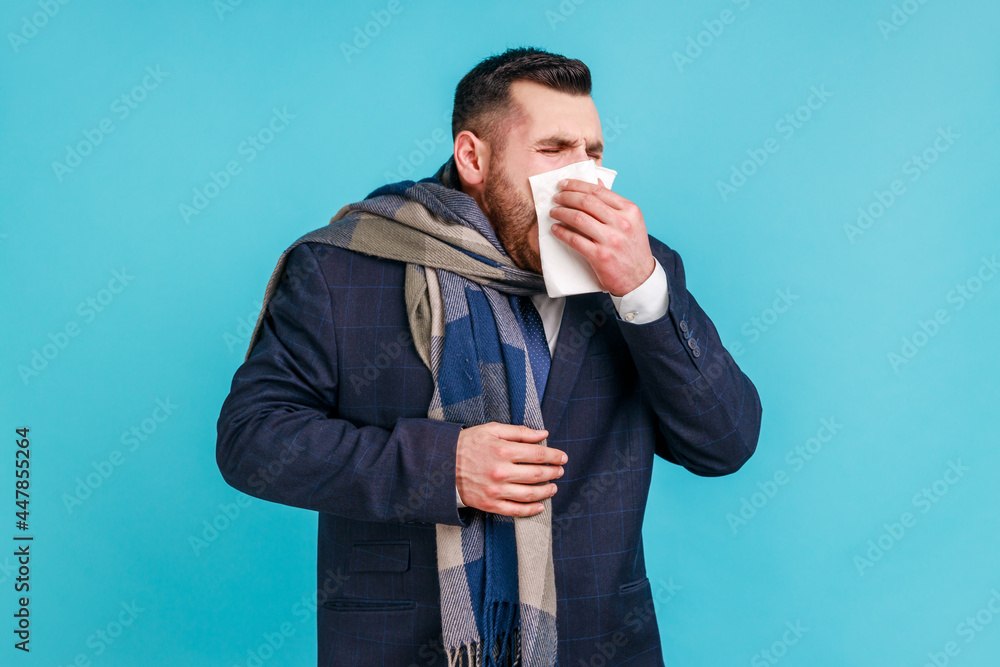 Cold and flu. Portrait of sick bearded young businessman in elegant suit standing, holding tissue on mouth and nose, being wrapped in warm scarf. Indoor studio shot isolated on blue background. Cold and flu. Portrait of sick bearded young businessman in elegant suit standing, holding tissue on mouth and nose, being wrapped in warm scarf. Indoor studio shot isolated on blue background.