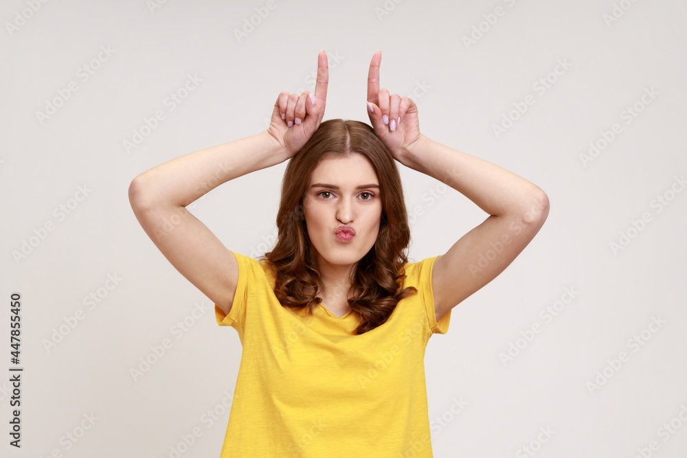 Bully teenager girl in yellow casual T-shirt looking threatening and showing bull horn sign, holding fingers on her head, conflicting person. Indoor studio shot isolated on gray background.