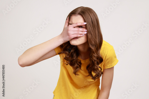 Portrait of beautiful teenager girl wearing yellow casual T-shirt spying, hiding and peeping through fingers, looking for secrets, curiosity. Indoor studio shot isolated on gray background.