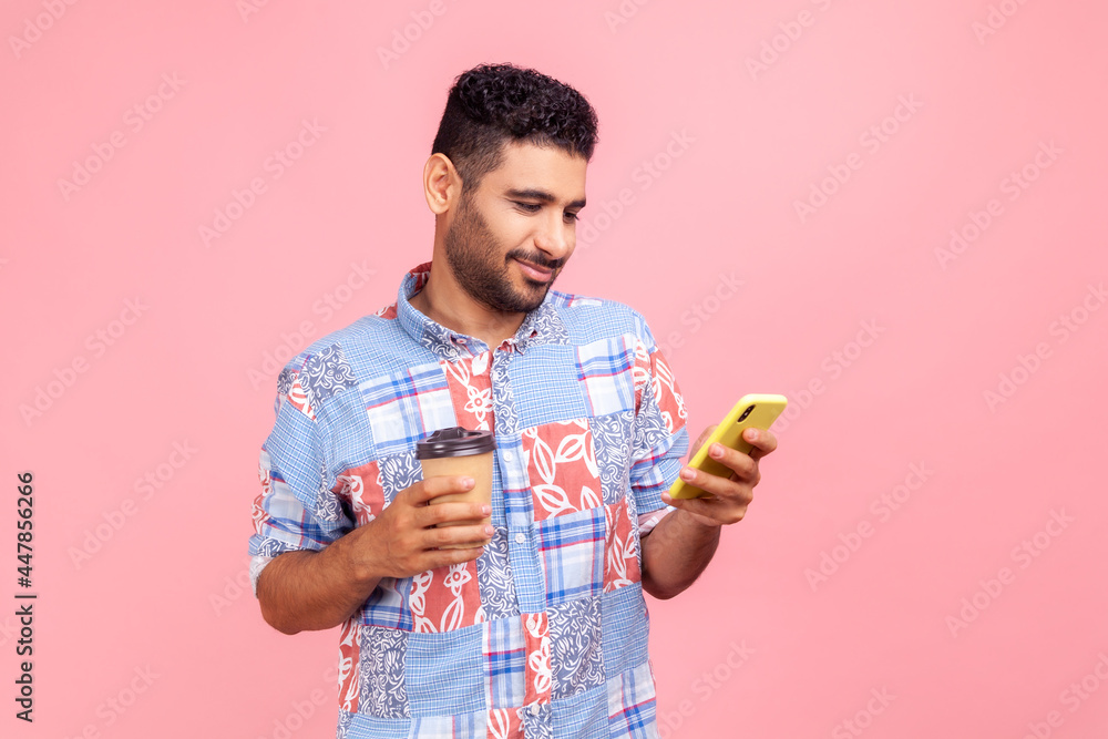 Portrait of satisfied handsome bearded man in blue casual style shirt standing with coffee and chatting on the phone, expressing positive emotions. Indoor studio shirt isolated on pink background. Portrait of satisfied handsome bearded man in blue casual style shirt standing with coffee and chatting on the phone, expressing positive emotions. Indoor studio shirt isolated on pink background.