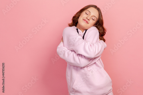 Portrait of selfish narcissistic brunette teen female in hoodie hugging herself and smiling with dreamy satisfied expression, love myself gesture. Indoor studio shot isolated on pink background © khosrork
