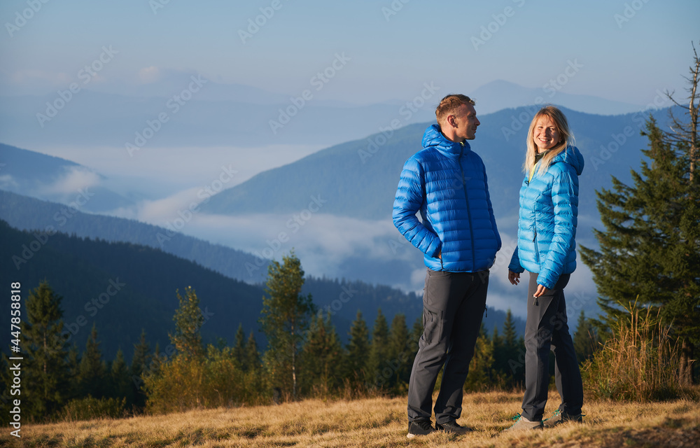 Couple of hikers standing with fun smile on grassy meadow in autumn on sunny day against the backdrop of mountain panorama. Hiking in the mountain spaces. Concept of travelling and relationships.