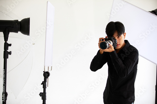 Asian young handsome successful male photographer wears black long sleeve shirt and jeans hold big DSLR cam in hands aiming forward to camera in indoor photography studio full of light and reflector