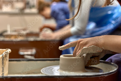 Hands of a ceramist while she is working on a potter's wheel against the background of other people working with clay