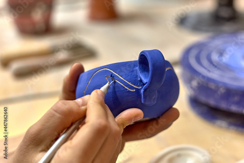 Obraz na plátně Hands of a ceramist who cuts a drawing with a knife on a painted blue cup in a w
