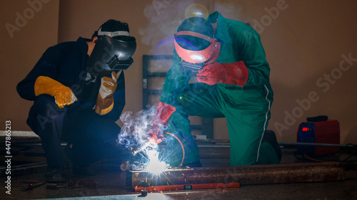 two engineers mechanics sitting and working in a workshop of a factory. They are helping each other to weld a piece of metal rod with a welding machine at night time