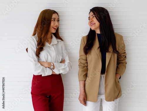 Portrait shot of two young Asian office workers in formal clothes standing, crossed arm and looking at each other and talking with a cute smiling on white background. Close friends and team concept.