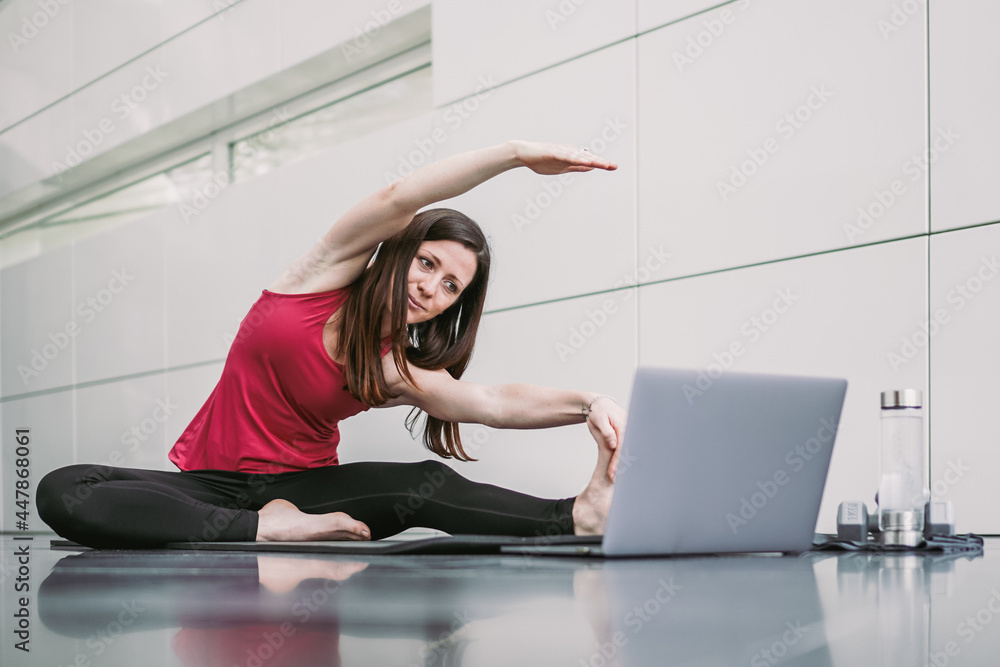 Fototapeta premium Young athletic fitness woman watching online leg stretching training video on yoga mat infront of a laptop at home studio indoor with panoramic window on reflective modern background 
