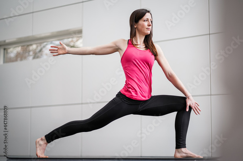  trained brunette woman in black leggin stretching slim body while practicing warrior 2 yoga pose indoor in a white modern studio on a mat 