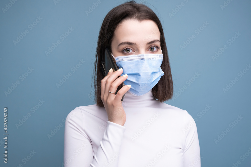 A young woman in a medical mask holds a mobile phone in her hands and makes a selfie on a blue background in the studio. A young woman in a medical mask holds a mobile phone in her hands and makes a selfie on a blue background in the studio.