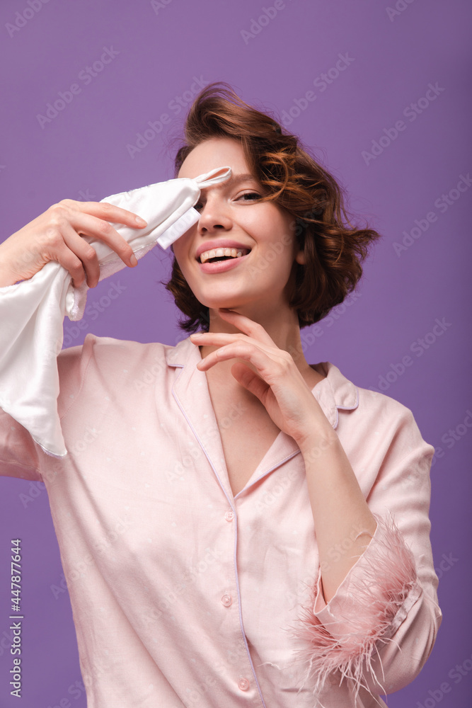 Beaty and skincare procedures. Cute young lady with dark curly hair and silk pink loungewear, making cleansing procedures, wiping white towel, isolated over violet background Beaty and skincare procedures. Cute young lady with dark curly hair and silk pink loungewear, making cleansing procedures, wiping white towel, isolated over violet background