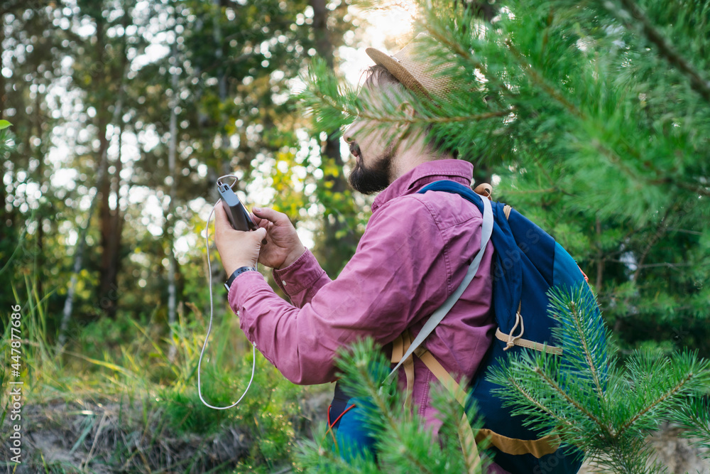 Tourist holds a smartphone with a portable charger in his hands. Man with a Power Bank charges the phone against the backdrop of nature and forest. Tourist holds a smartphone with a portable charger in his hands. Man with a Power Bank charges the phone against the backdrop of nature and forest.