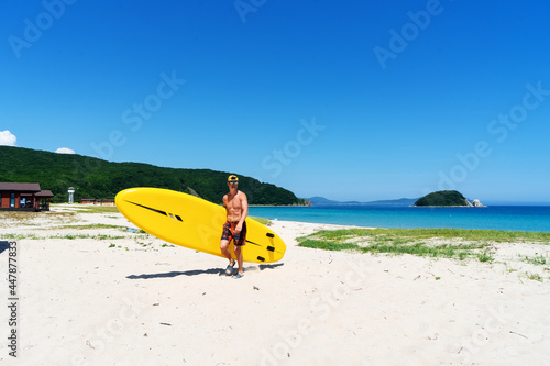 A man stands on a sandy shore and holds a large yellow paddle board.
