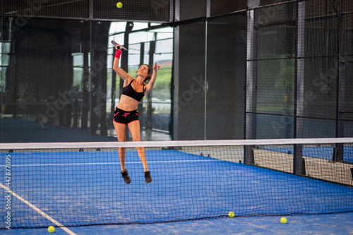 Woman playing padel in a blue grass padel court indoor - Young sporty woman padel player hitting ball with a racket © damianobuffo