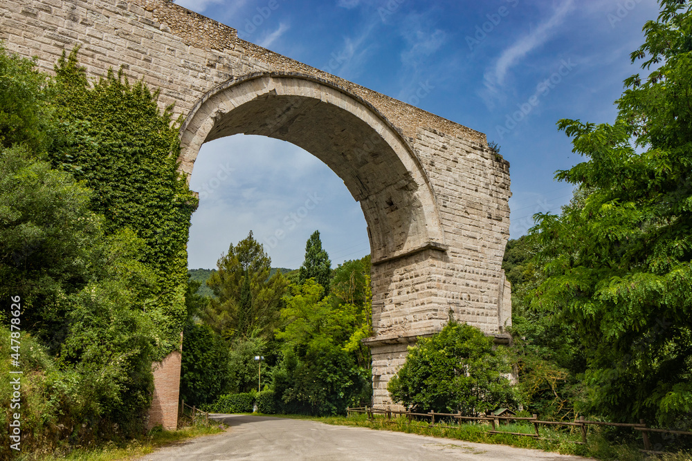 The ruins of the Roman arch bridge of Augustus, in Narni, Terni, Umbria. The remains of the bridge over the Nera river. The big and ancient stone arch, against the blue sky. Trees and dense vegetation