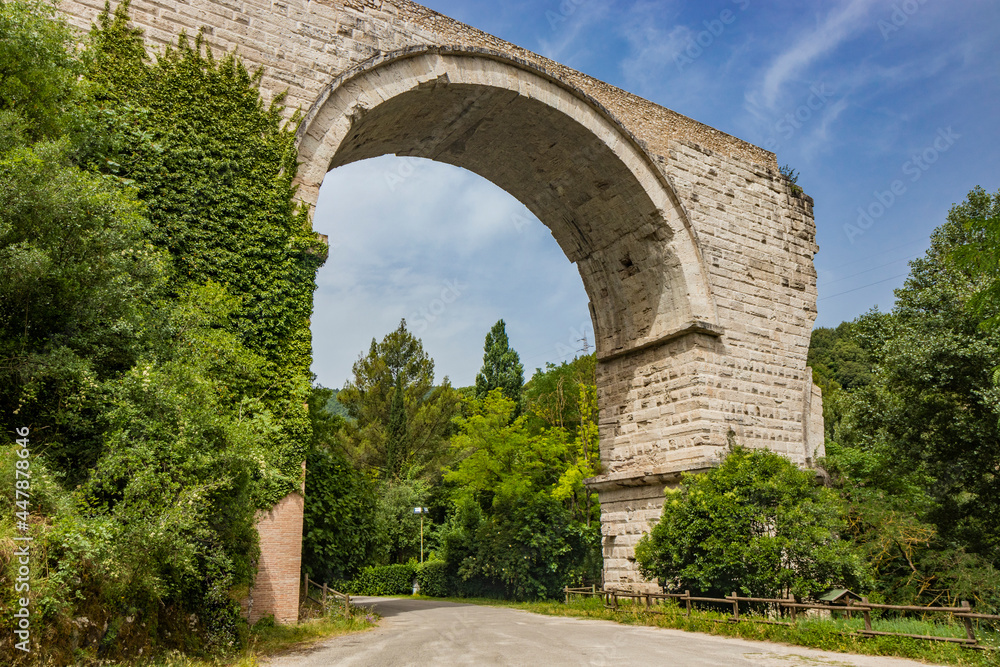 The ruins of the Roman arch bridge of Augustus, in Narni, Terni, Umbria. The remains of the bridge over the Nera river. The big and ancient stone arch, against the blue sky. Trees and dense vegetation