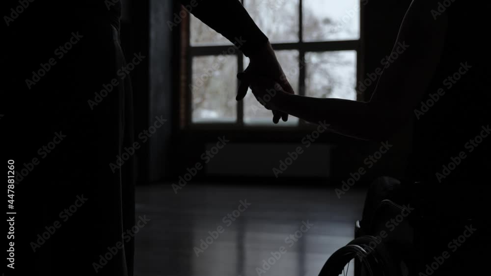 Close-up of hand in hand on background of window. Silhouettes of dance partners, man and disabled woman on wheelchair giving each other hand standing in the middle of gloomy dance hall Close-up of hand in hand on background of window. Silhouettes of dance partners, man and disabled woman on wheelchair giving each other hand standing in the middle of gloomy dance hall