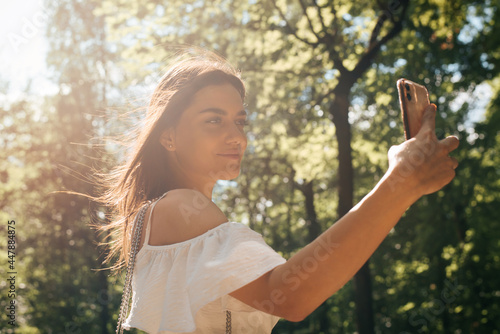 Attractive young brunette woman in a white summer dress making a positive selfie on mobile phone on a sunny summer day in the park. Smiling woman with hair fluttering in the wind outdoors, lifestyle