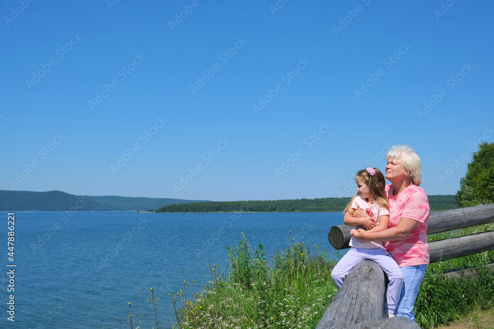 Grandmother and granddaughter stand together hugging and enjoy by the lake or sea. Summer vacation near the water. Having good times with grandparent outdoors Grandmother and granddaughter stand together hugging and enjoy by the lake or sea. Summer vacation near the water. Having good times with grandparent outdoors