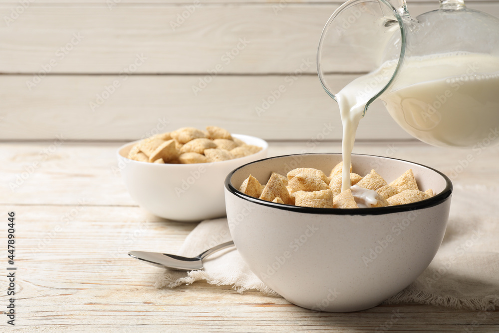 Pouring milk into bowl of tasty corn pads served on white wooden table. Space for text Pouring milk into bowl of tasty corn pads served on white wooden table. Space for text