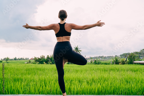 Back view of sportive girl dressed in black tracksuit standing in tree pose and feeling harmony mindfulness, slim fit girl enjoying morning hatha training near Asian rice fields in Ubud region
