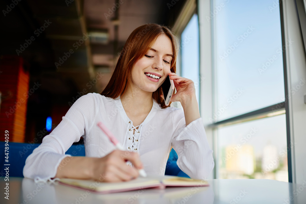Cheerful female smiling while writing informative notes during positive cellphone conversation for discussing organisation ideas, happy Caucasian student using mobile connection for call phoning Cheerful female smiling while writing informative notes during positive cellphone conversation for discussing organisation ideas, happy Caucasian student using mobile connection for call phoning
