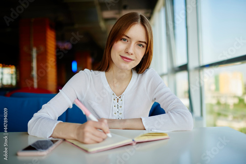 Portrait of young Caucaisan student with education textbook and pen for learning looking at camera, attractive youthful woman with notepad for studying and doing homework posing at table desktop