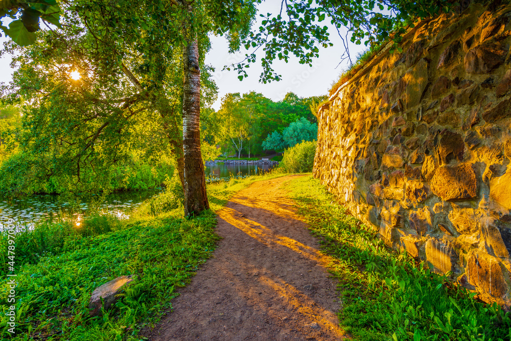 Naklejka premium Alley going between the fortress and the river. The sun shines through the foliage of the trees. Beautiful nature. Priozersk, Russia. View from the road near the fortress wall.