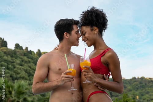 Multiracial young couple holding fruit juice glasses tenderly touching their foreheads. Surrounded by nature and trees on background.