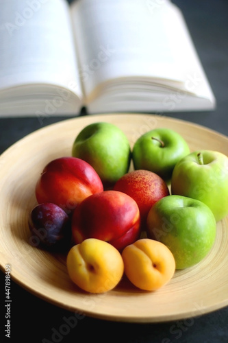 Wooden bowl with various summer fruit and open book on a table. Selective focus.