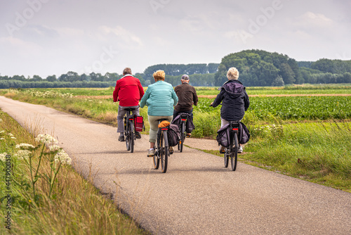 Two elderly unidentified couples with e-bikes cycle on a Dutch country road between fields in the province of North Brabant. The photo was taken on a slightly cloudy day in the summer season.