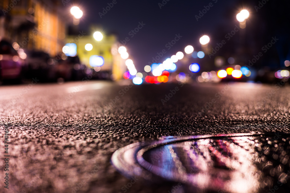 Rainy night in the big city, the headlights of the approaching cars on the road. Close up view of a hatch at the level of the asphalt
