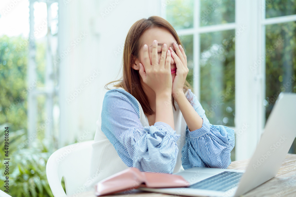 An Asian woman in a white shirt sits in a bakery, clutching her head in stress with her purse and laptop next to it. An Asian woman in a white shirt sits in a bakery, clutching her head in stress with her purse and laptop next to it.